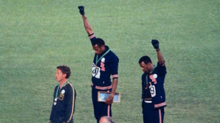 Gold medalist Tommie Smith (center) and bronze medalist John Carlos (right) showing the raised fist on the podium after the 200 m race at the 1968 Summer Olympics; both wear Olympic Project for Human Rights badges. Peter Norman (silver medalist, left) from Australia also wears an OPHR badge in solidarity with Smith and Carlos. Angelo Cozzi (CC0)